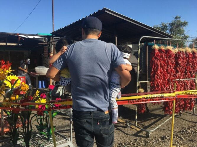 man with his back to the camera carrying two children with ristras in the background